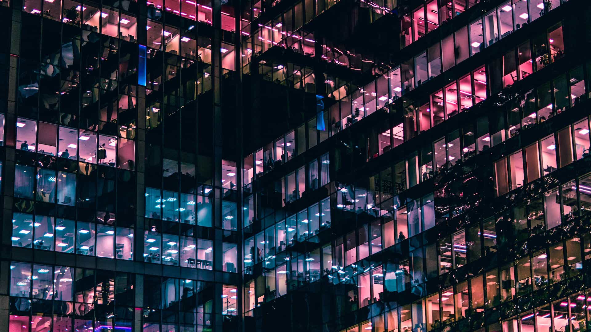 Night view of a modern office building with lit windows showing interior workspaces in blue and pink lighting.
