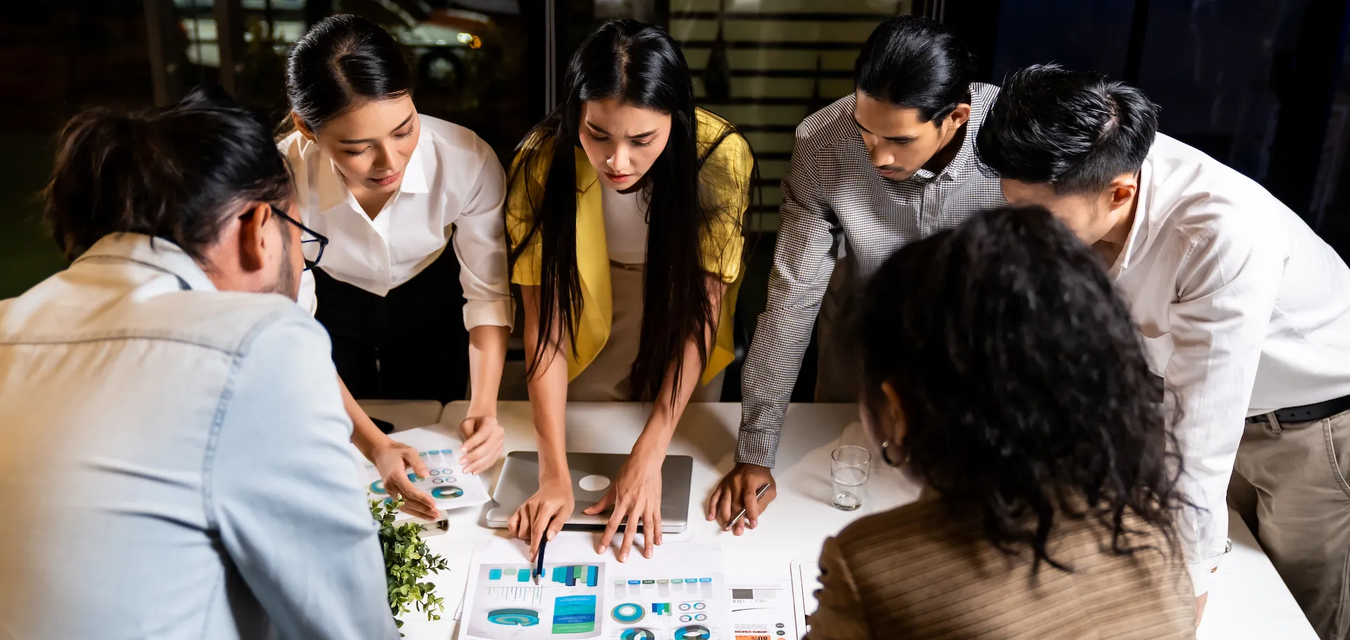 Group of six diverse colleagues leaning over a table reviewing colorful charts and graphs during a collaborative meeting.