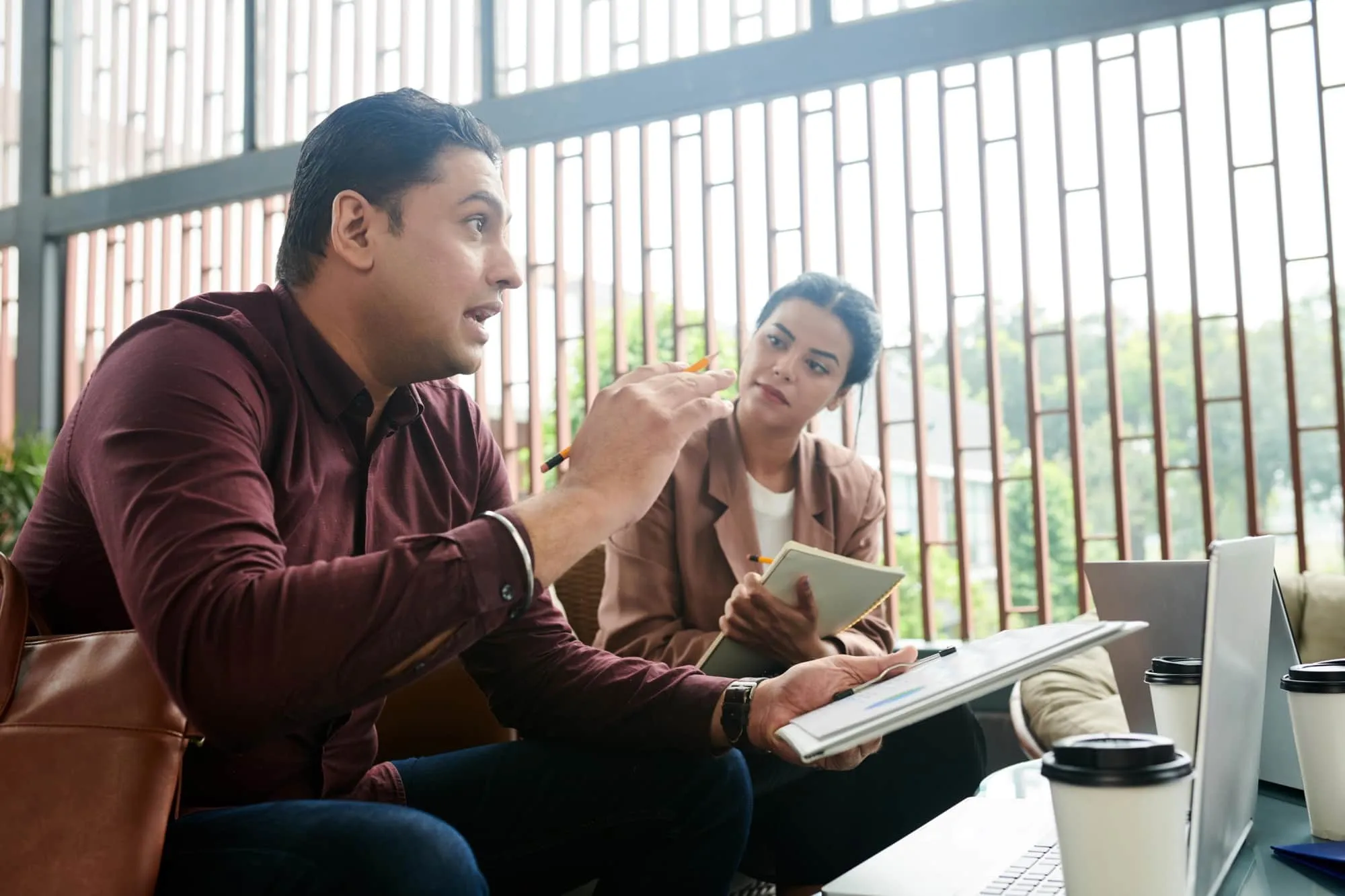 Two professionals discussing work with documents and laptops in an office with large windows.