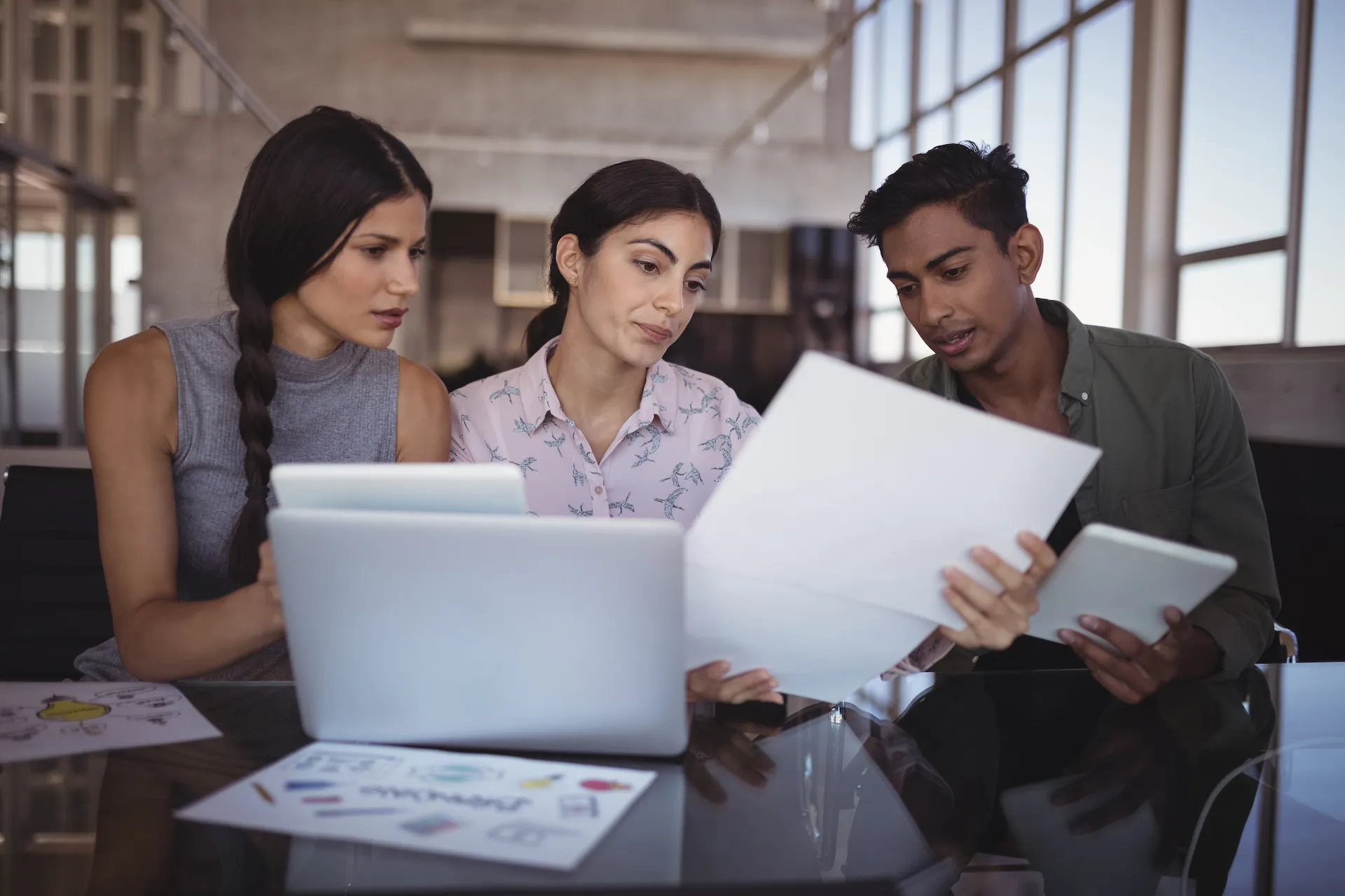 Three young professionals reviewing documents and working on digital devices in a modern office.
