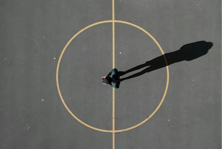 A person standing alone at the center of a basketball court casting a long shadow.