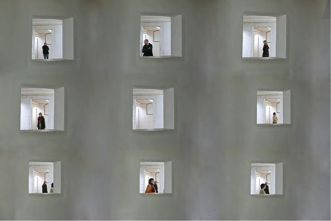 Nine square windows on a grey wall, each showing people standing or walking inside a white corridor.