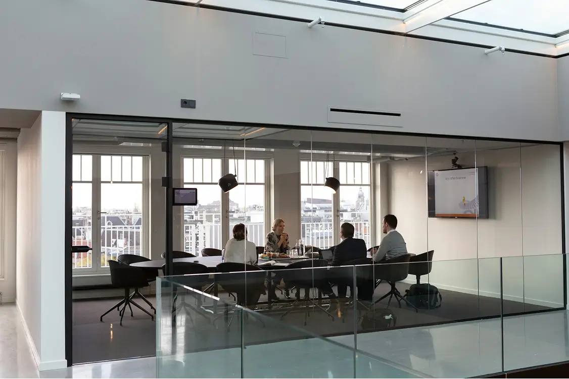 Four people seated around a table in a modern glass-walled office meeting room with large windows overlooking a cityscape.