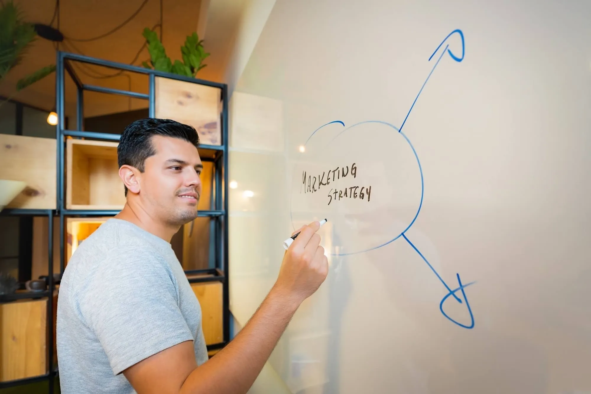 Man in gray shirt writing 'Marketing Strategy' on a glass board with arrows pointing away.